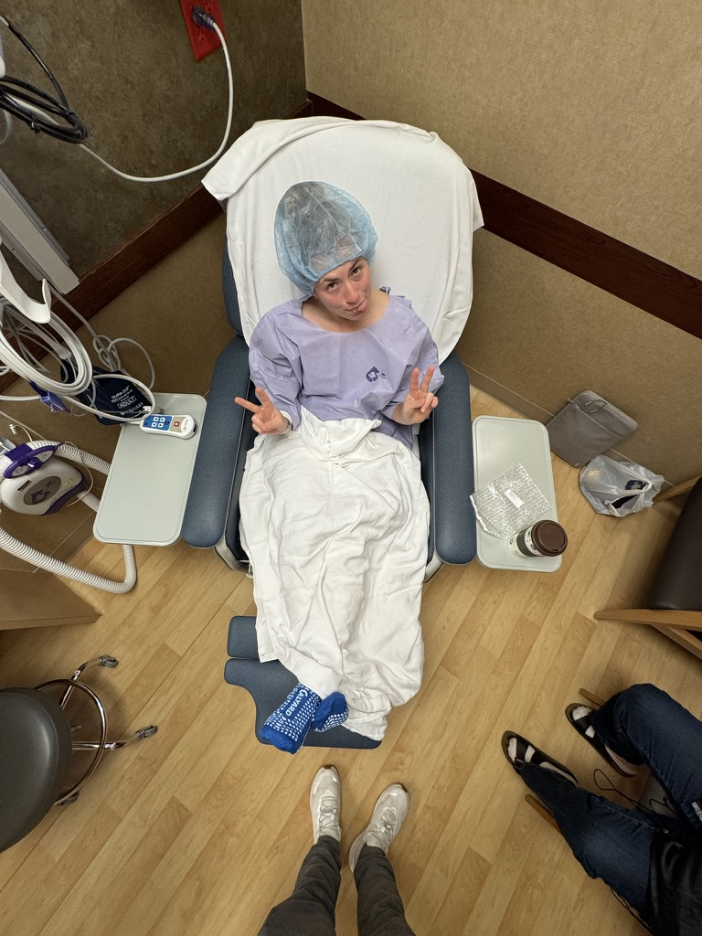 Person in hospital gown and cap seated in recliner chair covered with blanket making peace signs.